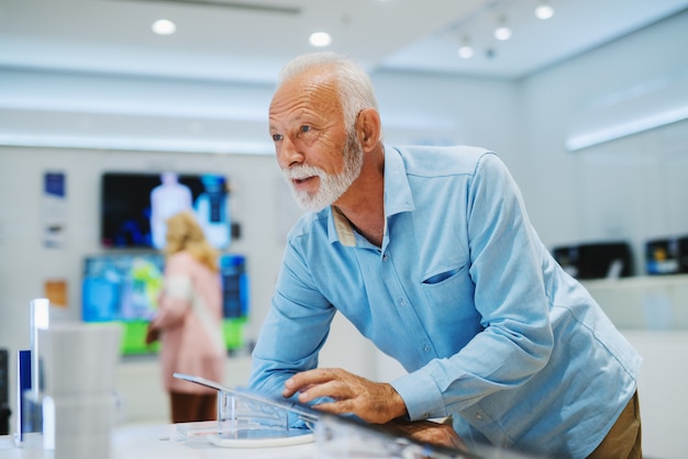 Senior man with smiling face testing tablet he want to buy and leaning on the stand while standing in tech store.