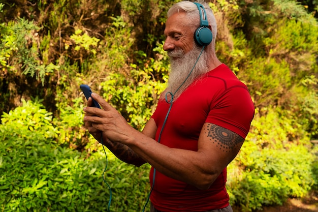 Senior man with white beard doing running outdoor on the nature, holding phone and choosing music