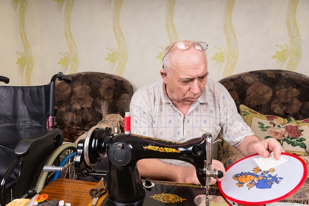 Senior Man Working On Needle Point Wall Hanging and Seated in front of Old Fashioned Manual Sewing Machine in Living Room at Home