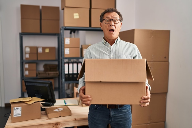 Senior man working at small business ecommerce holding carboard bx in shock face looking skeptical and sarcastic surprised with open mouth