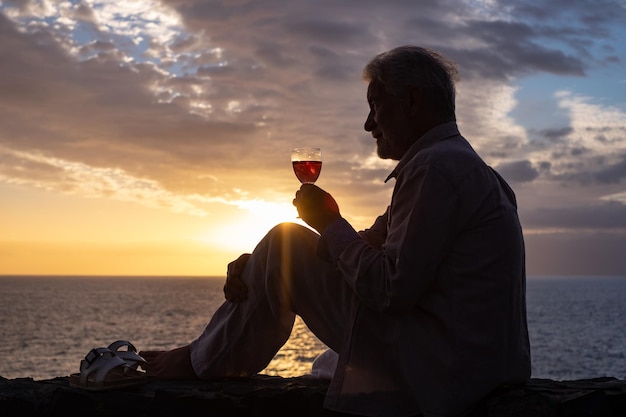 Senior man zit voor de zee bij zonsondergang met een glas wijn horizon over water Positief gevoel voor oudere man die geniet van vakantie of pensioen