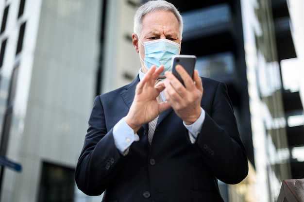 コロナウイルスのパンデミックから保護するためにマスクを着用しながら屋外でスマートフォンを使用するシニアマネージャー