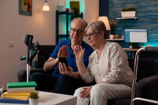 Пожилые пожилые люди, состоящие в браке, используют технологию видеозвонка для онлайн-конференции на смартфоне дома. Пенсионеры разговаривают с семьей с помощью удаленного интернет-общения, сидя на диване
