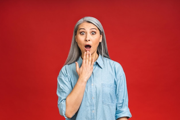 Senior mature aged pretty woman feeling shocked and excited laughing amazed and happy because of an unexpected surprise isolated over red background