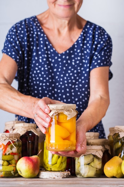 Senior mature woman holding in hands jar with homemade preserved and fermented food. Variety of pickled and marinated vegetables, fruit compote. Housekeeping, home economics, harvest preservation