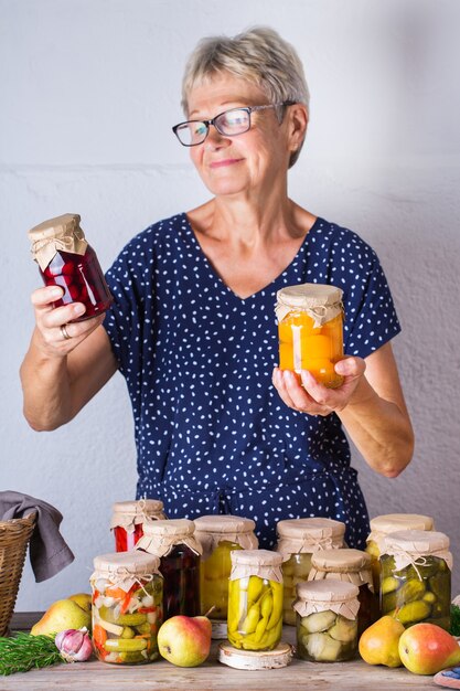 Senior mature woman holding in hands jar with homemade preserved and fermented food. Variety of pickled and marinated vegetables, fruit compote. Housekeeping, home economics, harvest preservation