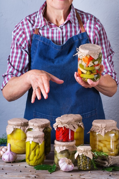 Senior mature woman holding in hands jar with homemade preserved and fermented food. Variety of pickled and marinated vegetables. Housekeeping, home economics, harvest preservation