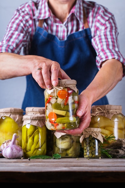 Senior mature woman holding in hands jar with homemade preserved and fermented food. Variety of pickled and marinated vegetables. Housekeeping, home economics, harvest preservation