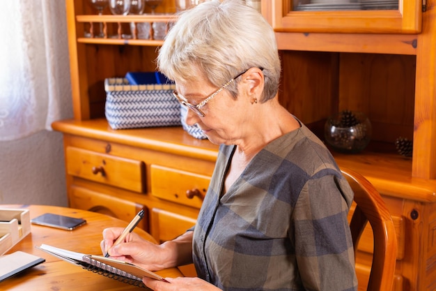 Senior mature woman making notes while sitting near the table