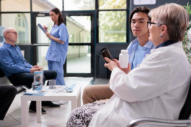 Senior medic explaining medication treatment to asian patient\
showing painkiller pills during checkup visit consultation in\
hospital room. diverse people standing in lobby waiting to start\
examination