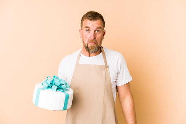 Senior met een cake geïsoleerd op beige muur haalt zijn schouders op en opent verwarde ogen.