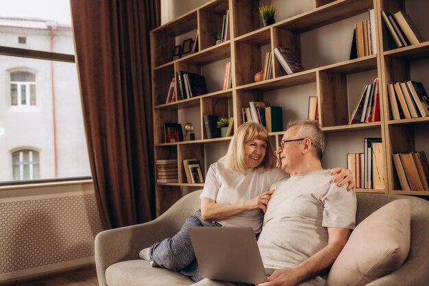 Senior middle aged happy couple embracing using laptop together smiling elderly family reading news shopping online at home older people and computer or good vision after laser correction concept