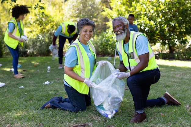 Photo senior mixed race couple spending time outside with their family, presenting a garbage bag with garbage in it, looking at the camera and smiling, all wearing blue volunteers t shirts, on a sunny day