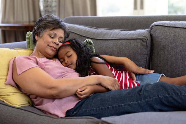 Senior mixed race woman enjoying her time at home, lying on a couch, embracing her granddaughter, with their eyes closed