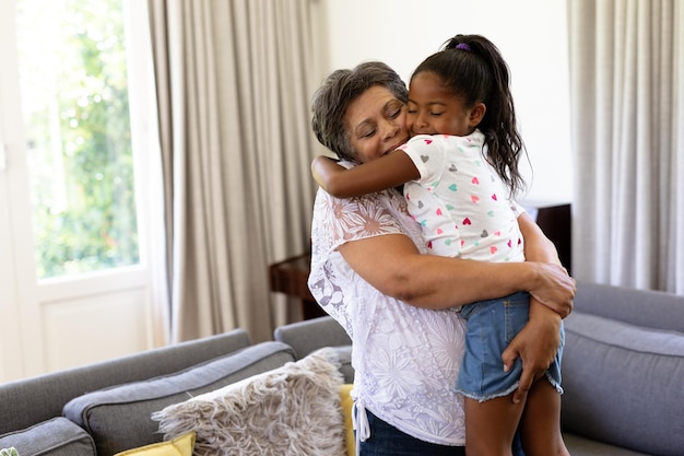 Senior mixed race woman and her granddaughter enjoying their time at home together, standing by a couch, woman is holding girl up, embracing and smiling