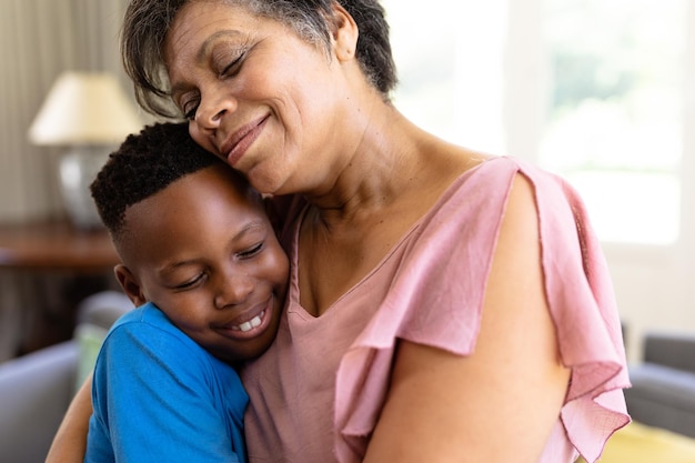 Senior mixed race woman and her grandson enjoying their time at home together, sitting on a couch, embracing and smiling with their eyes closed