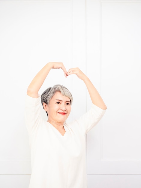 Senior mooie grijsharige vrouw met een casual shirt glimlachend in liefde die de vorm van een hartsymbool met handen doet. Romantisch begrip.