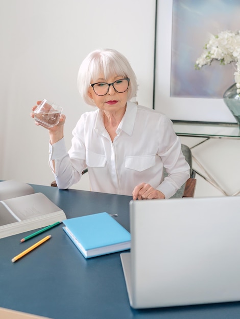 Senior mooie grijze haren vrouw in witte blouse drinkwater tijdens het werk op kantoor