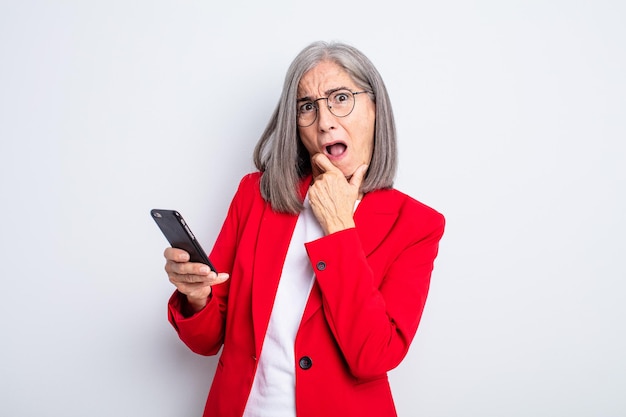 Senior mooie vrouw met mond en ogen wijd open en hand op kin... bedrijfs- en telefoonconcept