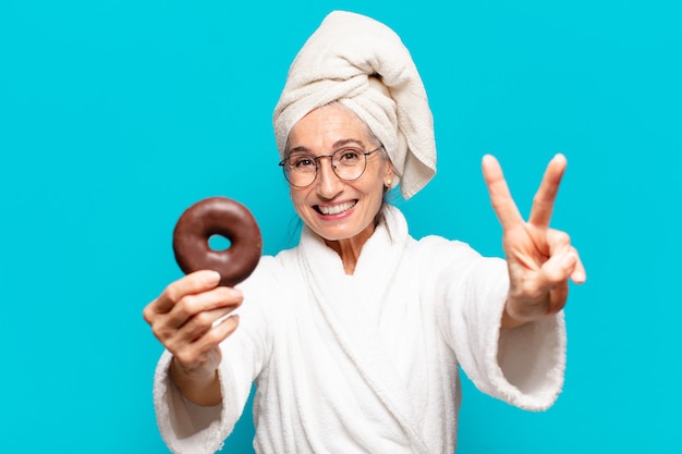 Senior mooie vrouw na het douchen en het dragen van badjas en ontbijten met een donut