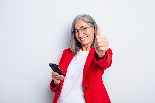 Senior mooie vrouw voelt zich trots, positief glimlachend met duimen omhoog... zakelijk en telefoonconcept