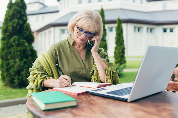 Senior mooie zakelijke vrouw zittend op een stoel en het maken van aantekeningen in een notitieblok met een telefoon