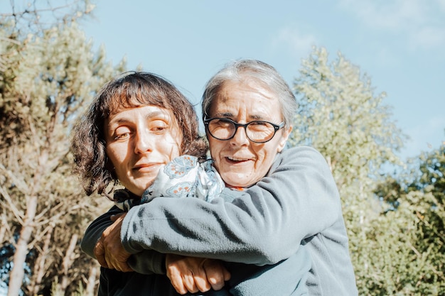 Senior mother embracing middle age daughter smiling in a park during a walk trecking exercise. Exercising together helping our elder.