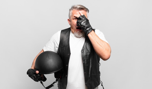 Senior motorbike rider looking shocked, scared or terrified, covering face with hand and peeking between fingers