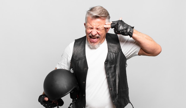 Senior motorbike rider looking unhappy and stressed, suicide gesture making gun sign with hand, pointing to head