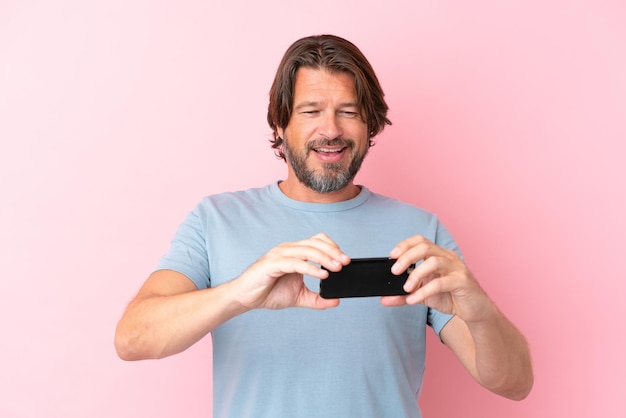 Senior Nederlandse man geïsoleerd op roze achtergrond spelen met de mobiele telefoon