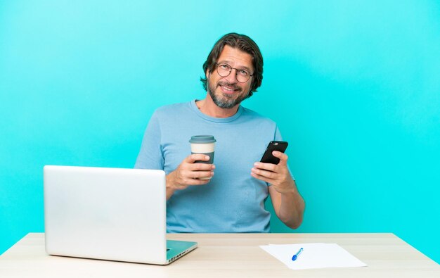 Senior Nederlandse man in een tafel met een laptop geïsoleerd op blauwe achtergrond met koffie om mee te nemen en een mobiel