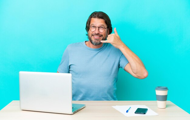 Senior Nederlandse man in een tafel met een laptop geïsoleerd op blauwe achtergrond telefoon gebaar maken. Bel me terug teken