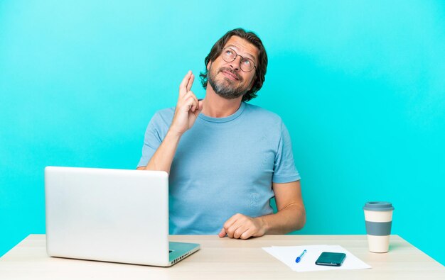 Senior Nederlandse man in een tafel met een laptop geïsoleerd op een blauwe achtergrond met vingers die kruisen en het beste wensen