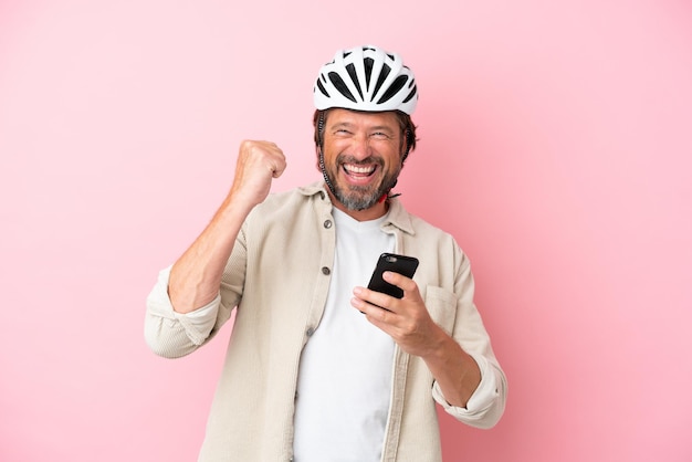 Senior Nederlandse man met fietshelm geïsoleerd op roze achtergrond met telefoon in overwinningspositie