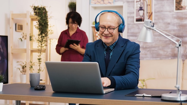 Senior old man listening music on headphones while working on laptop in living room. Wife using tablet in the background.