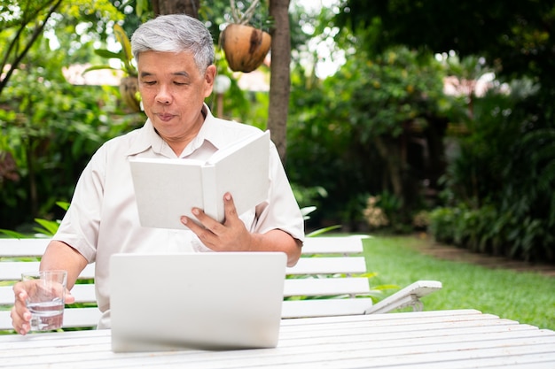 senior old man reading a book in the park, drinking water and using his laptop