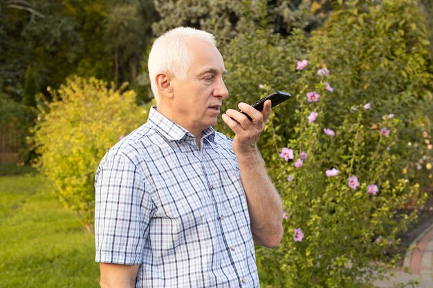 Senior oude man met behulp van mobiele telefoon met gesproken zoekopdrachten in groen park, moderne technologieën