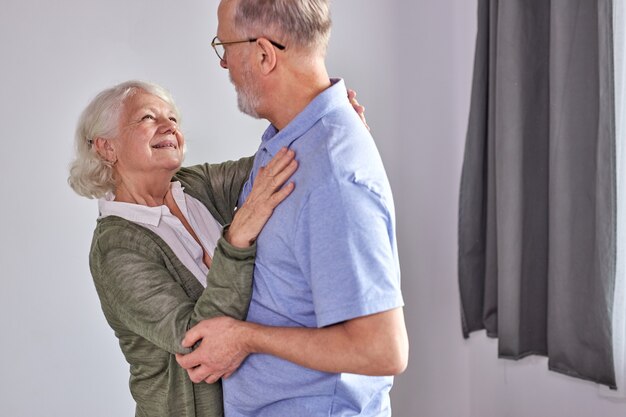 Senior paar dansen in de woonkamer, man met de hand van volwassen vrouw samen genieten van plezier, vakantie, vrije tijd pensioen ifestyle thuis doorbrengen. in vrijetijdskleding