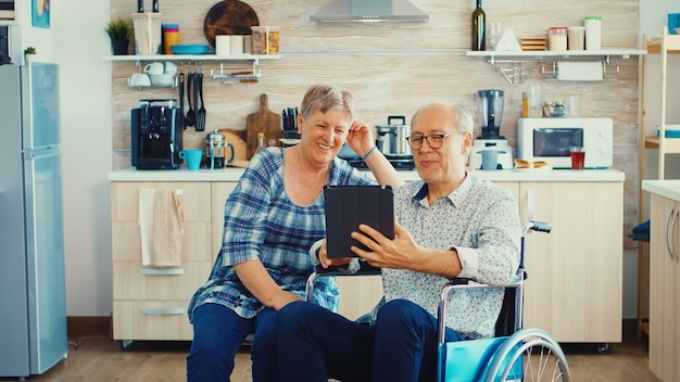 Senior paar lachen en zwaaien tijdens een videogesprek met kleinkinderen met behulp van tablet pc in de keuken. Verlamde gehandicapte oude bejaarde man met behulp van moderne communicatietechnologie.