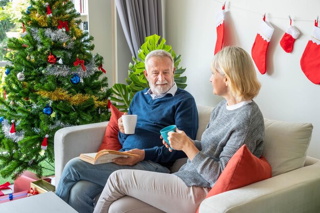 Senior paar man en vrouw lezen een boek en drinken koffie thee zittend op de bank met kerstversiering op de achtergrond