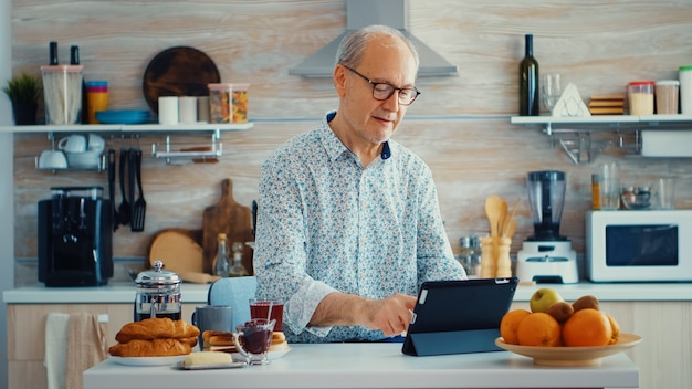 健康的なライフスタイルを持っている朝食時にキッチンでタブレットPCを閲覧しているシニアの親。モバイルアプリ、現代のインターネットオンライン情報を使用して定年のタブレットポータブルパッドPCを持つ高齢者