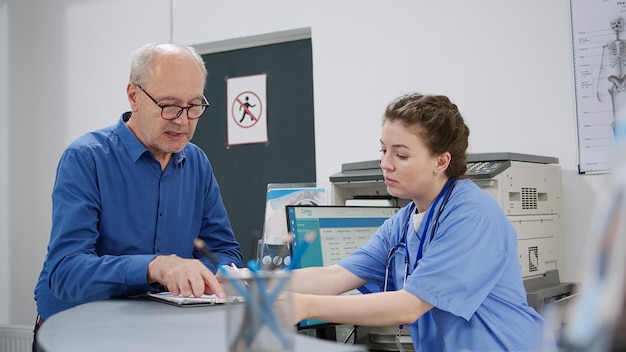 Senior patiënt in gesprek met verpleegkundige bij receptie in gezondheidscentrum, met medische consultatie afspraak. oude man die controlerapporten invult om een zorgverzekering te ontvangen. uit de hand geschoten.