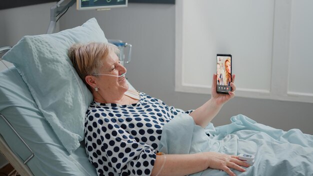 Senior patient talking to family on video call in hospital ward
bed. woman with sickness using smartphone and online video
conference for remote communication with niece and daughter.