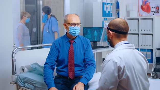 Senior patient wearing mask at doctor appointment during COVID 19. Modern private hospital or clinic. Healthcare medical physician consultation. Retired man consulting with healthcare worker