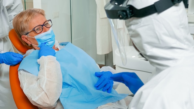 Senior patient with dental bib and protection mask talking with doctor in coverall about dental problem in stomatological clinic during coronavirus pandemic. Dentist wearing face shield and gloves