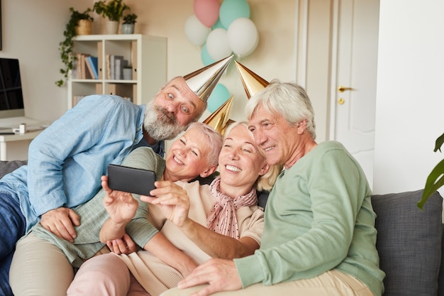 Photo senior people in party hats sitting on sofa and posing at camera together they making selfie portrait on mobile phone