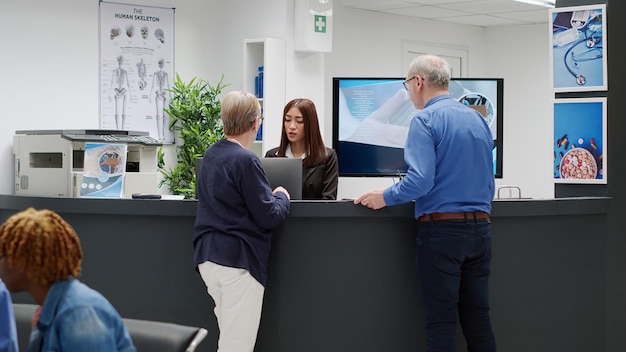 Senior people talking to receptionist at hospital reception
desk, asking about medical appointment and healthcare system. old
patients sitting at reception counter in waiting room lobby.