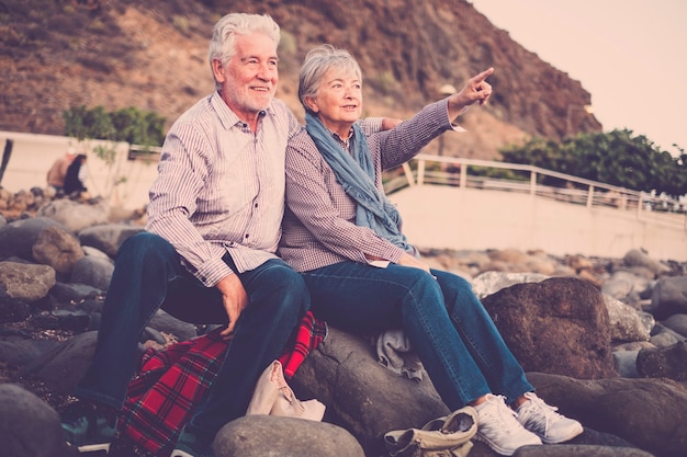 写真 一緒に恋をしている高齢者-夕焼けの地平線に向かって腕を伸ばして抱きしめ、見て、指差しながらビーチに座っている老夫婦。休暇、余暇、リラクゼーションの概念