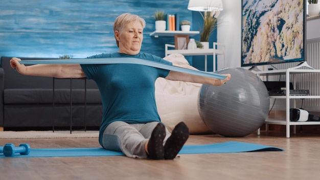 Senior persoon die weerstandsband trekt om op yogamat te oefenen. oudere vrouw die een elastische riem gebruikt om thuis een training te doen, waarbij de armspieren worden uitgerekt. oude volwassene trainen met apparatuur