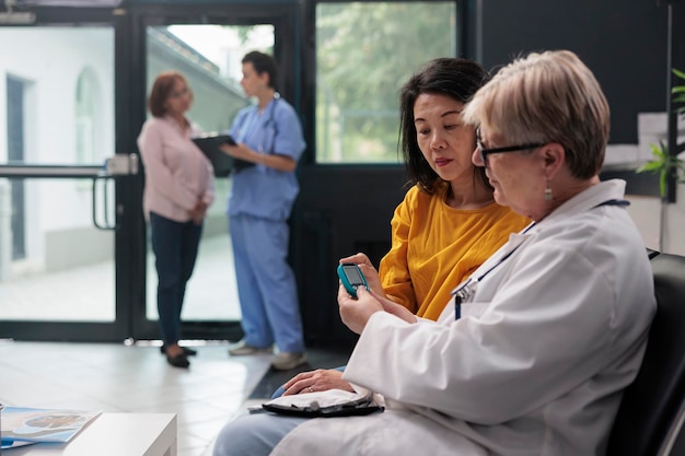 Senior physician consulting asian patient with glucometer,\
measuring insulin and glucose level from blood sample. medic and\
woman doing diabetic control at checkup visit examination.